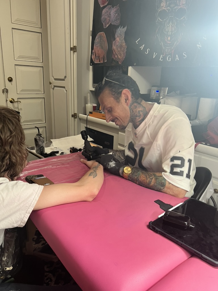Joshua Cole tattoos black arrow on inner forearm at pink table with Vegas banner behind — Joshua…, Work of Art Tattoo & Piercing, Las Vegas