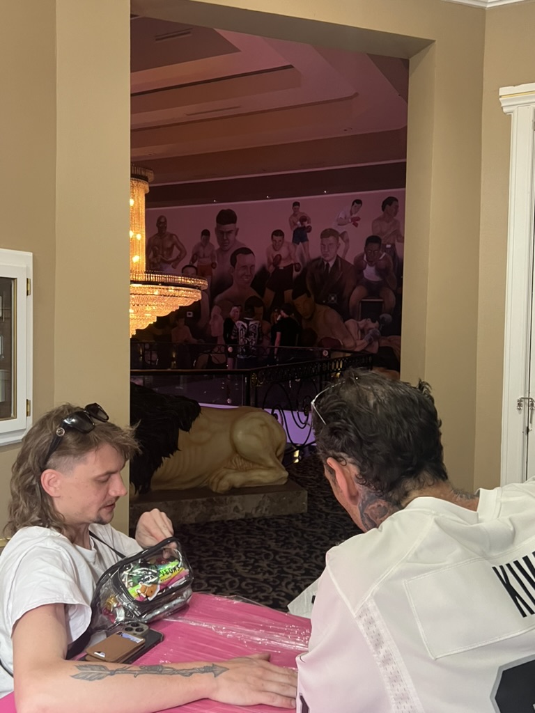 Men sit at pink offsite table; client shows fresh arrow forearm tattoo near lion statue — Joshua…, Work of Art Tattoo & Piercing, Las Vegas