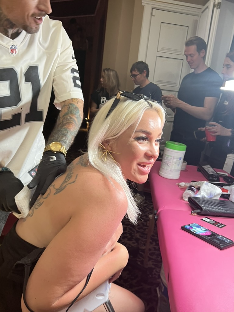Joshua Cole wipes fresh back tattoo on smiling blonde woman at crowded offsite setup — Joshua…, Work of Art Tattoo & Piercing, Las Vegas