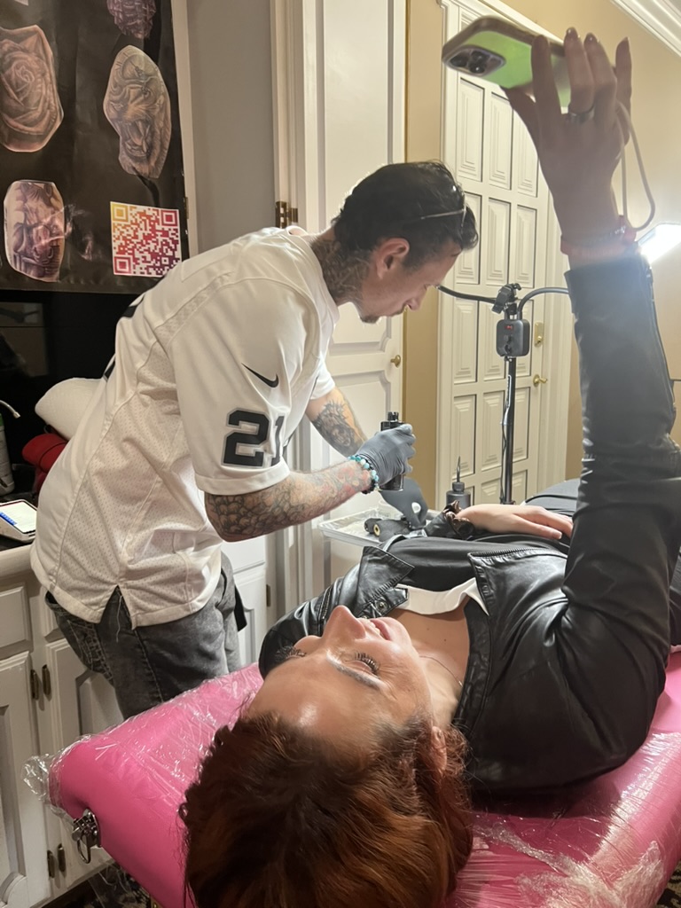 Client on pink table films Joshua Cole tattooing her arm with phone selfie — Joshua Cole offsite…, Work of Art Tattoo & Piercing, Las Vegas