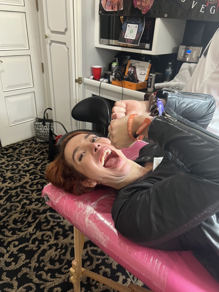Red-haired client laughs on pink-wrap table wearing orange Stacks wristbands — Joshua Cole…, Work of Art Tattoo & Piercing, Las Vegas