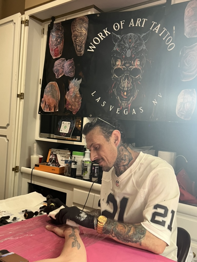 Joshua Cole tattoos forearm at pink table beneath Work of Art Las Vegas skull banner — Joshua…, Work of Art Tattoo & Piercing, Las Vegas