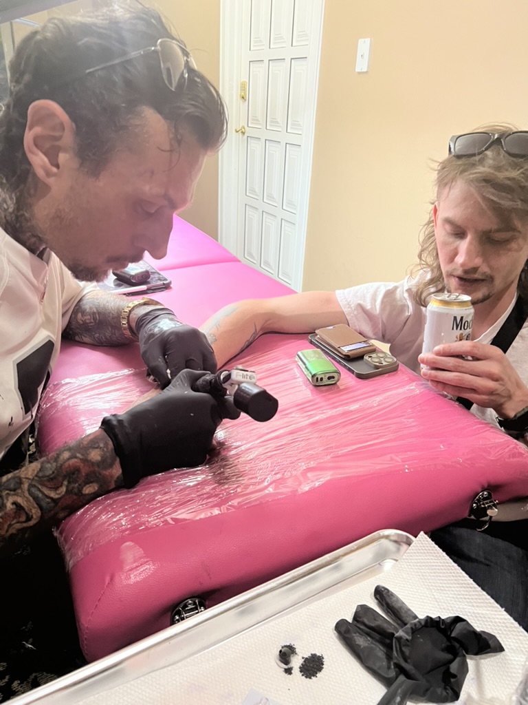 Joshua Cole tattoos forearm as mullet client holds Modelo on pink table — Joshua Cole offsite…, Work of Art Tattoo & Piercing, Las Vegas