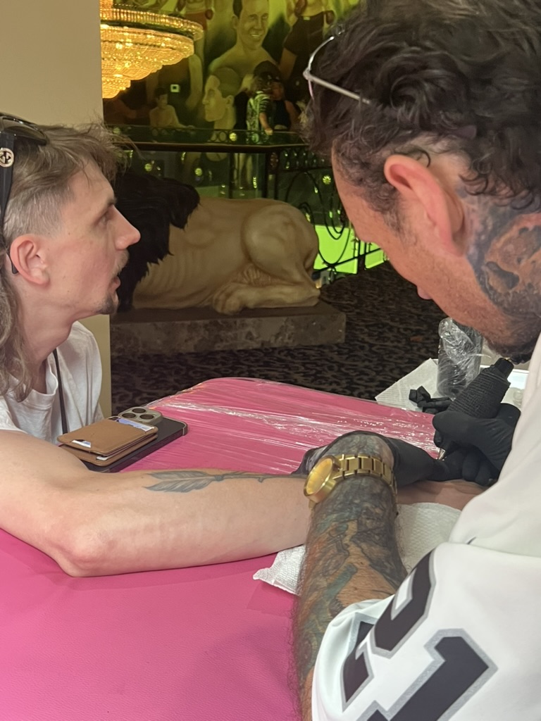 Joshua Cole tattoos forearm on pink table beside lion statue and boxing mural offsite — Joshua…, Work of Art Tattoo & Piercing, Las Vegas