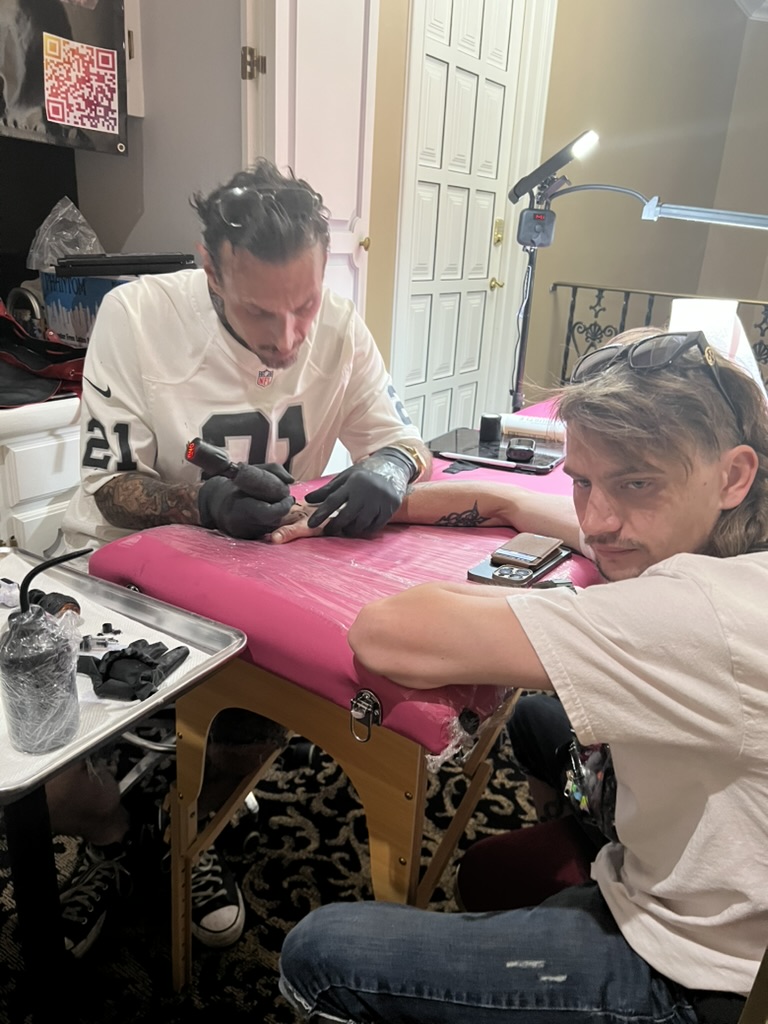 Joshua Cole tattoos client's hand on pink table; mullet client faces camera — Joshua Cole…, Work of Art Tattoo & Piercing, Las Vegas