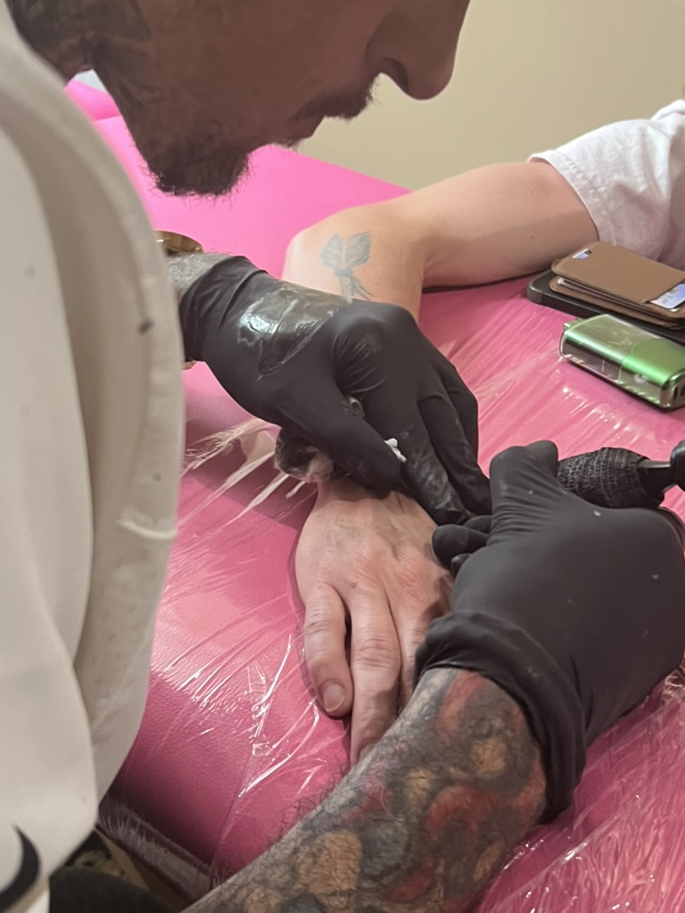 Joshua Cole tattoos back of client's hand on pink-wrapped table; arrow on forearm — Joshua Cole…, Work of Art Tattoo & Piercing, Las Vegas