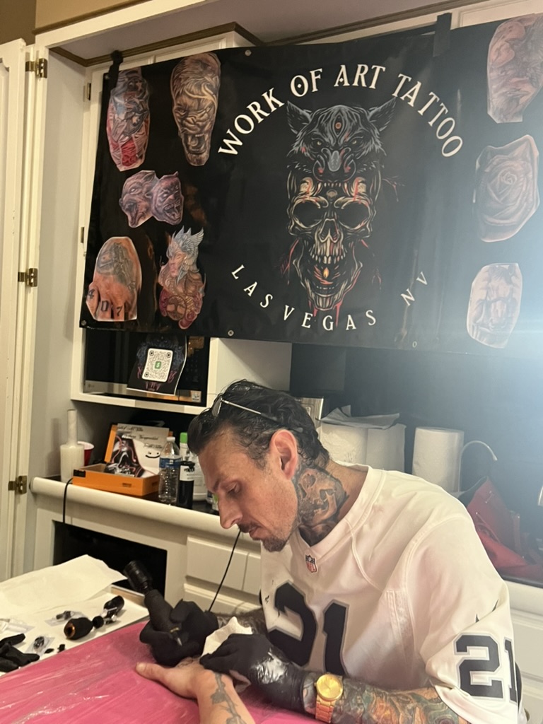 Joshua Cole tattoos client's hand on pink table beneath Work of Art Las Vegas banner — Joshua…, Work of Art Tattoo & Piercing, Las Vegas
