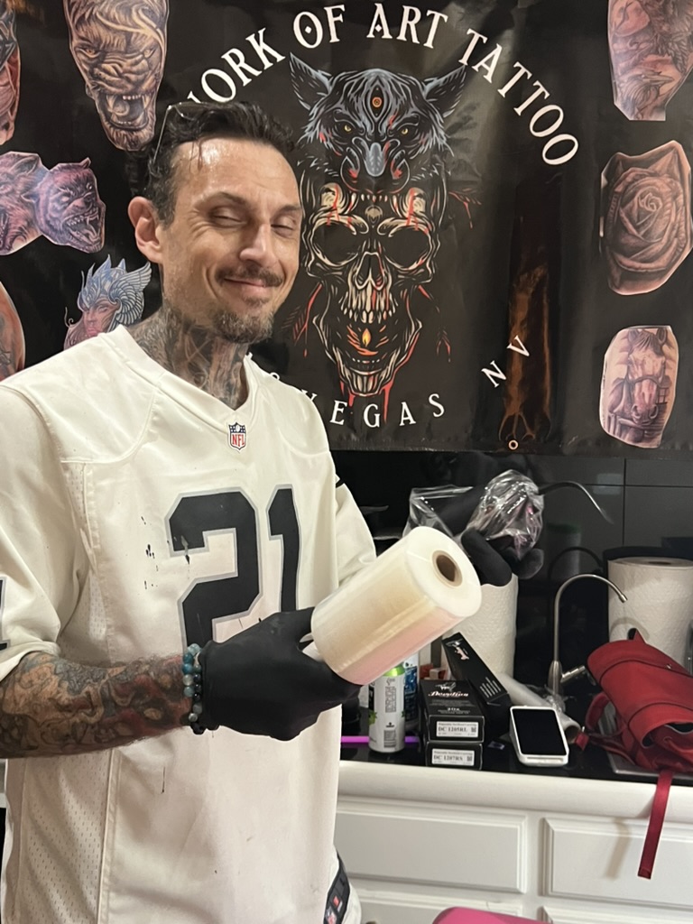 Joshua Cole smiles holding barrier film and wrapped machine at WOA banner — Joshua Cole offsite…, Work of Art Tattoo & Piercing, Las Vegas