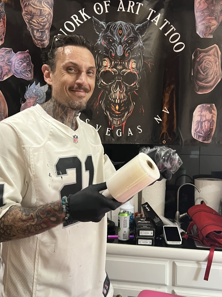 Joshua Cole grins holding clear barrier film roll at mobile banner — Joshua Cole offsite…, Work of Art Tattoo & Piercing, Las Vegas