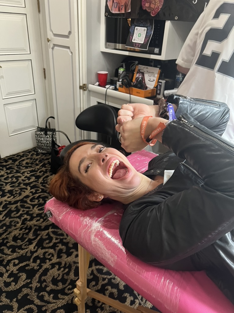 Client grins wide on pink tattoo table with festival wristbands at VIP party — Joshua Cole…, Work of Art Tattoo & Piercing, Las Vegas