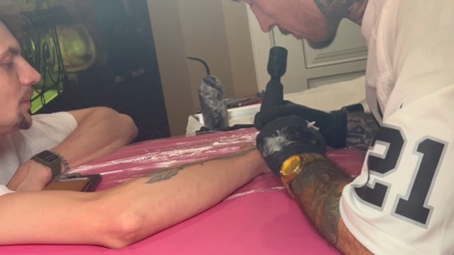 Raiders jersey artist tattoos rose on forearm at pink-covered offsite table — Joshua Cole…, Work of Art Tattoo & Piercing, Las Vegas