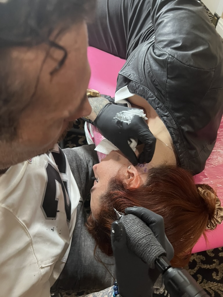 Joshua Cole tattoos purple script behind client's ear on pink-wrapped offsite station — Joshua…, Work of Art Tattoo & Piercing, Las Vegas