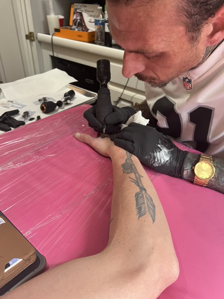 Joshua Cole tattoos small design on hand beside arrow-and-rose forearm at pink table — Joshua…, Work of Art Tattoo & Piercing, Las Vegas