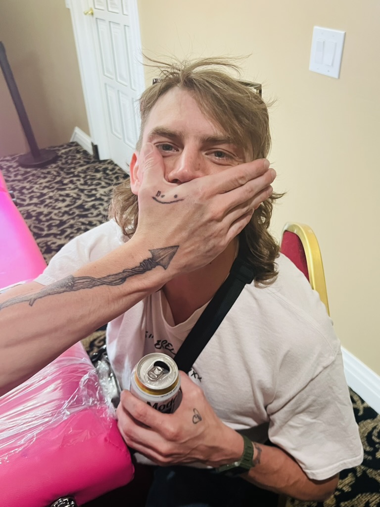 Smiley hand tattoo covers man's mouth beside spear forearm tattoo at pink offsite table — Joshua…, Work of Art Tattoo & Piercing, Las Vegas