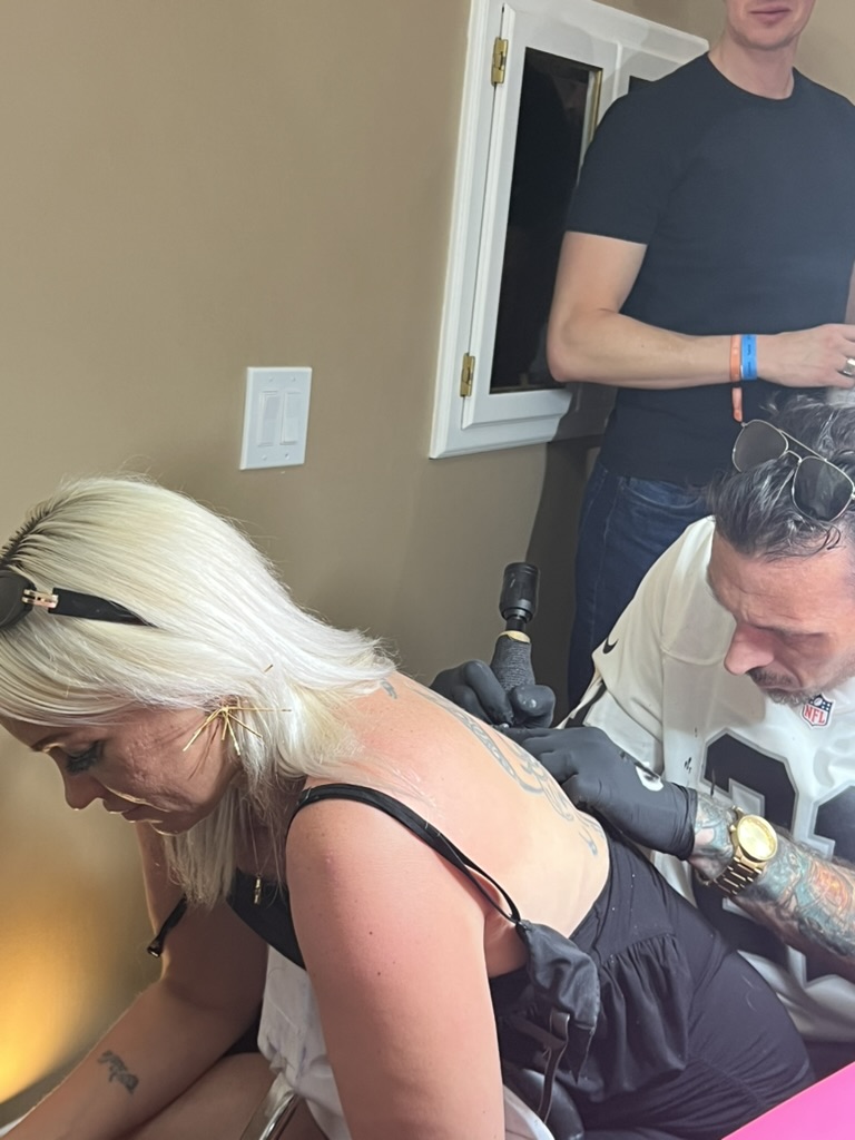 Joshua Cole tattoos upper-back piece on blonde client in Raiders jersey setup — Joshua Cole…, Work of Art Tattoo & Piercing, Las Vegas