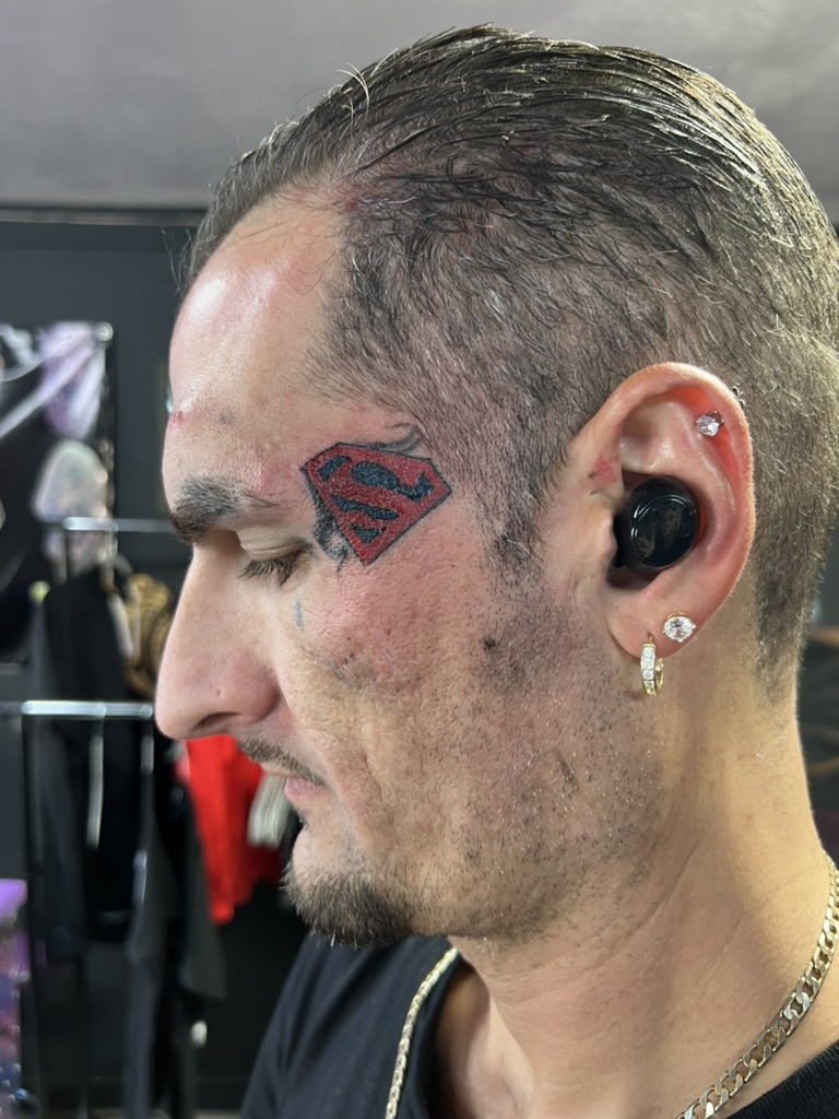 Color Superman S shield with smoke accents on left temple near eyebrow. — Joshua Cole, Work of…, Work of Art Tattoo & Piercing, Las Vegas