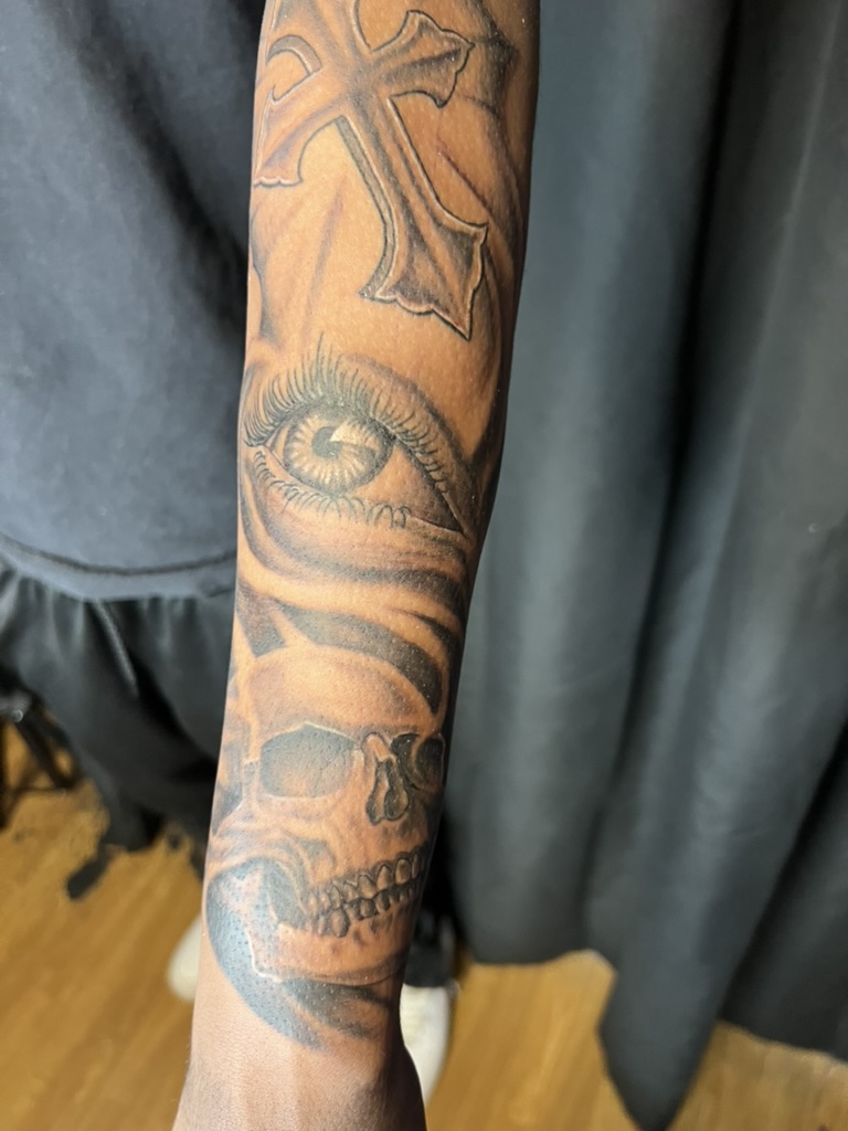 Black and grey cross, eye, and skull forearm tattoo on brown skin. — Joshua Cole, Work of Art…, Work of Art Tattoo & Piercing, Las Vegas