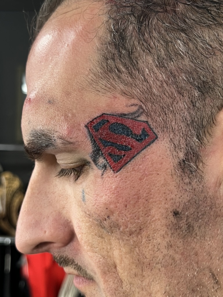 Red and black Superman S shield tattoo on a man's left temple. — Joshua Cole, Work of Art Las…, Work of Art Tattoo & Piercing, Las Vegas